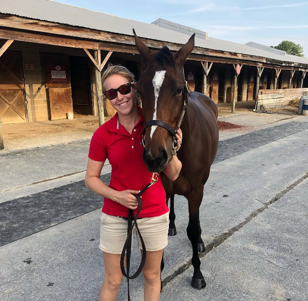 A smiling woman in a red polo standing beside a bay horse in a barn aisle on a summer evening.